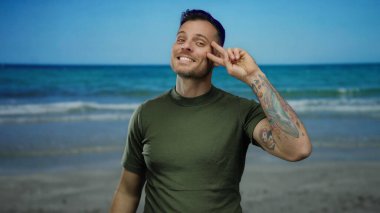 Dövmeli gülümseyen adam deniz kenarında barış işareti yapıyor. Arka planda okyanus var. Güneşli gökyüzünün altında kumsal gününün tadını çıkarıyor..