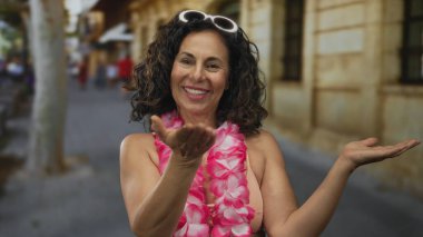 Orta yaşlı İspanyol bikinili kadın ve Hawaii zambağı açık avuçlarını bir şehir caddesinde sunar; tropik tatil neşesi neşesi.