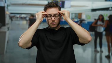 Kapalı bir havaalanı terminalinde gözlük takan sakallı genç adam hareketli bir ortamda gezginlerle çevrili modern bir seyahat sahnesi yaratıyor..