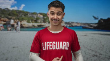Kırmızı tişörtlü genç İspanyol cankurtaran açık mavi deniz ve arka planda yeşillikle güneşli sahilde güvenle dikiliyor..