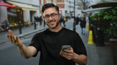 Sakallı genç adam canlı şehir caddesinde telefonu tutarken kameraya el hareketi yapıyor. Kendine güveni tam ve açık havada hareketli bir ortam yaratıyor..