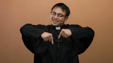 Kahverengi arka planda el kol hareketi yaparken gülümseyen ve işaret eden gözlüklü bir rahip, dostça bir ifade ve dini kıyafet sergiliyor.