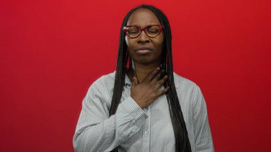 Uzun örgülü bir kadın, kırmızı bir duvara yaslanmış gözlükleriyle kendini kötü hissediyor. Alnına ve yanağına dokunarak rahatsızlığını dile getiriyor..