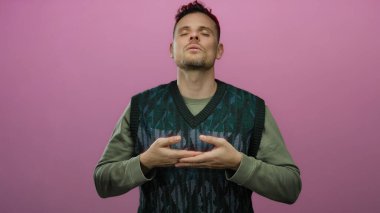 Genç İspanyol bir adam, yeşil bir süveter giyerken sakin ve odaklanmış bir şekilde, izole edilmiş pembe bir arka plan önünde meditasyon yapıyor.