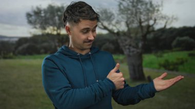 Serin bir park ortamında avucunun içini gösterip el hareketi yapan genç İspanyol bir adam..