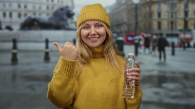 Sarı kazaklı ve bereli bir kadın su şişesi tutuyor. Arka planda bulanık bir parmak izi var. Açık şehir ortamında kendine güveni ve günlük tarzı var..