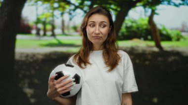 Güneşli park ortamında elinde futbol topu tutan bir kadın yemyeşil bir ortamda neşeli bir ifade sergiliyor. Dışarıda kaygısız bir boş zamanı yansıtıyor..