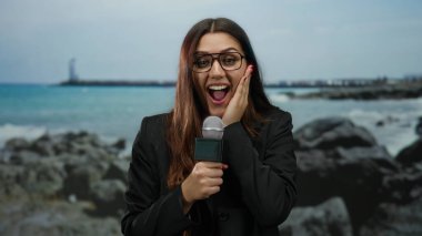 Sahilde muhabir olarak mikrofon tutan bir kadın gözlüklü açık havada okyanus manzarası karşısında şaşırmış görünüyor..