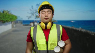 Elinde planlar tutan genç bir adam deniz kenarında sakin bir deniz altyapısı olan güvenlik ekipmanları giyiyor inşaat temaları ve açık havada mimari kavramlar sergiliyor..