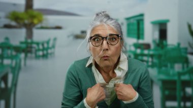 Gözlüklü yaşlı bir kadın, bir restoranın terasında, güneşli bir caddede, yeşil sandalyelerle çevrili bir yerde şaşkınlıkla kendini işaret ediyor..