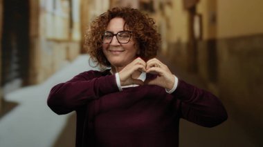 Kıvırcık saçlı ve gözlüklü bir kadın şehir arka planında gülümserken kalp hareketi yapıyor, dışarıda mutluluk ve pozitifliği ifade ediyor..