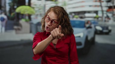 Kırmızı tişörtlü kadın gün boyunca şehir sokaklarında ifade ile işaret ediyor. Arka planda arabalar ve binalar olan kentsel bir ortamda yoğunluk taşıyor.