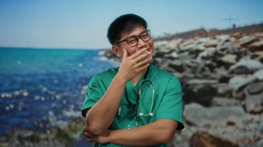 Gözlüklü ve steteskoplu genç adam kayalık bir plajda gülüyor mavi deniz arkaplanı ve berrak gökyüzü açık havada doğa ve sağlık hizmetlerini birleştiriyor..