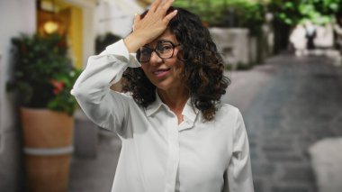 Beyaz tişörtlü, kıvırcık saçlı, şehir sokaklarında düşünceli görünen bir kadın..