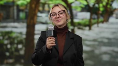 Parkta mikrofonu elinde tutan kadın muhabir, sanki güvenle gülümserken birini daha yakına davet ediyormuş gibi hareket ediyor..