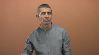 Gri gömlekli bir adam kahverengi bir duvara yaslanmış meraklı ve dikkatli bir şekilde bej arka planda dikkatle dinlerken