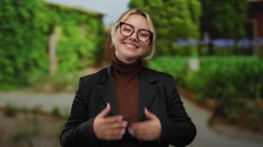 Açık hava parkı ortamında gülümseyen yeşil arka planda gözlük takan ve siyah ceketli bir kadın doğal bir ortamda pozitiflik ve rahatlama sergiliyor.