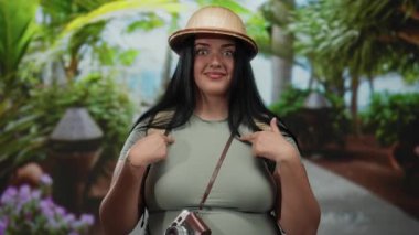 Şaşırmış ifadesi olan bir kadın turist, tropikal bir şapka takmış ve elinde antika bir kamerayla kendini açık hava parkında gösteriyor..