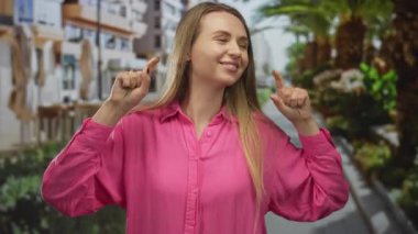 Pembe gömlekli kadın palmiye ağaçlarıyla dolu bir caddede neşe içinde dans ediyor. Canlı bir şehir bölgesinde, canlı bir enerjiyle mutluluğu ifade ediyor..