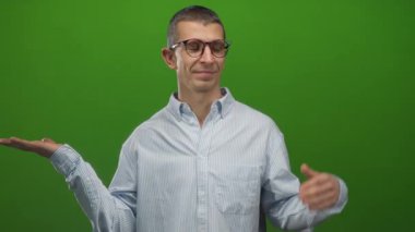 Yeşil arka plan duvarına yaslanmış gözlüklü bir adam, sorgulama jestinde elleri açık, çizgili bir gömlek giymiş, bir seçim ya da karar ima ediyor.