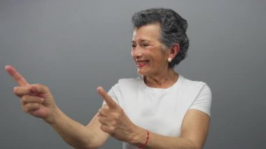 Gri saçlı, gülümseyen ve orta yaşlı bir kadın. Gri arkaplanlı modern, minimalist bir ortamda, günlük beyaz kıyafetler giyerken neşe ve pozitifliğini ifade ediyor..