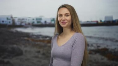 Sahilde gülümseyen bir kadın, gri süveter giyerek kıyı gününün tadını çıkarıyor. Rahatlamayı öneriyor..