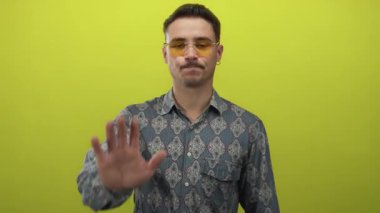 Güneş gözlüklü genç adam, parlak sarı arkaplan karşısında el işaretini uzatarak, izole bir ortamda uyarı veya reddetme mesajı iletiyor..