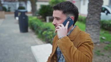 Akıllı telefondan konuşan, güneşli bir parkta açık havada kahverengi ceket giyen, modern şehir yaşam tarzını ve iletişimini sergileyen genç bir adam..