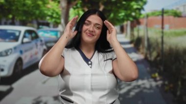 Şehir caddesinde genç, kıvrımlı bir kadın, neşeli bir şekilde gülümserken ve elleriyle yüzünü örtüyor, şehir manzarası ve güneşli gökyüzünün altında park edilmiş arabalarla çevrili..