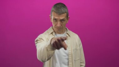 Kafkasyalı bir adam elini sallayarak birinin gitmesi için işaret eder, canlı pembe bir arka plana karşı durur, açık renk bir gömlek giyer ve doğrudan kameraya bakar..