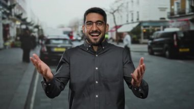 Gözlüklü gülümseyen genç adam kalabalık bir şehir sokağında dikiliyor açık havada neşe saçıyor..