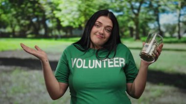 Kadın gönüllü elinde dolar dolu kavanozla güneşli açık hava parkında gülümsüyor.