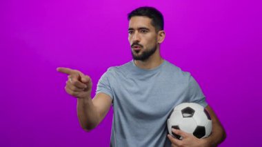 Genç adam futbol topuyla parlak pembe arka plana işaret ediyor, azmini gösteriyor ve sportif bir bağlamda odaklanıyor..