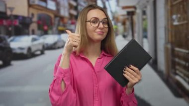 Pembe gömlekli bir kadın, elinde bir kitapla kentin işlek bir caddesinde kendine güvenen ve tarz sahibi bir şekilde dikiliyor..