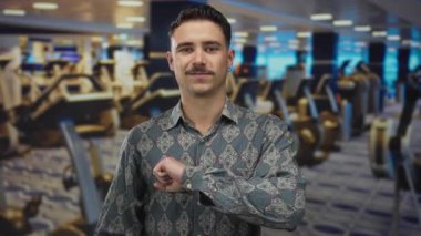 Genç İspanyol asıllı bir adam, spor salonunda, spor aletleriyle dolu, azimli ve odaklanmış bir şekilde, yumruğunu kameraya doğru uzatıyor..