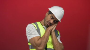 Beyaz bir şapka ve sarı yansıtıcı yelek giyen İspanyol genç adam kırmızı bir stüdyoda el ele bastırır; yorgunluk.