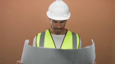 Adam kahverengi stüdyoda güvenlik yeleği ve beyaz şapka giyerken iki eliyle planı yuvarladı; stratejik planlama güveni.