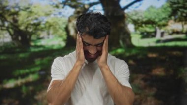 Beyaz tişörtlü adam, orman parkında elleri başının üstünde, benekli güneş ışığı altında geniş bir gülümseme içinde, sükunet;.