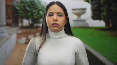 Kadın, açık havada sürpriz bir ifadeyle ellerini kaldırıyor. Bahçenin yanında Latin kökenli genç özellikler ve balıkçı yaka kazağının altında uzun saçlı bir çerçeve var..