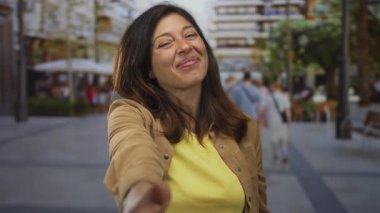 Sıcak gülüşlü, günlük kahverengi ceketli kadın hareketli caddede kameraya doğru elini uzatıyor; bağlantı.