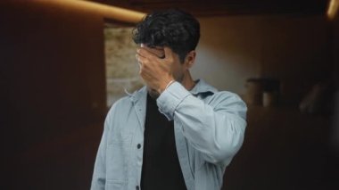 Kapalı bir stüdyo odasında eli ile yüzünü kapatan adam yumuşak sıcak aydınlatma ve ahşap panel duvarlar ile; endişe.