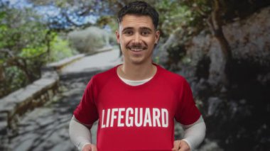 Parkta kırmızı bir cankurtaran tişörtü içinde gülümseyen genç İspanyol bir adam. Güneşli bir günde çevresi yeşillikle çevrili..