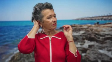 Kıdemli, kır saçlı, canlı kırmızı elbiseli bir kadın deniz kenarında manzaralı bir plaj geçmişine dokunuyor..