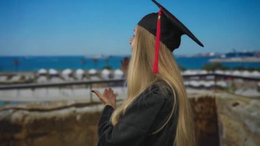 Mezuniyet şapkalı ve cüppeli bir kadın kameraya gülümsüyor arka planda manzaralı bir liman ve tekne, unutulmaz bir açık hava başarı anı yakalıyor..