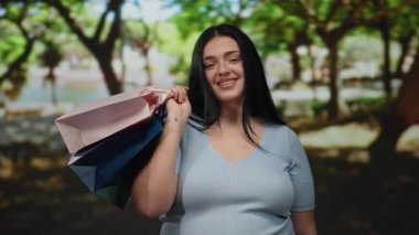 Parkta alışveriş yapmaktan hoşlanan bir kadın elinde renkli çantalarla arka planda ağaçlar ve açık havada mutluluğunu yansıtan mutlu bir gülümseme.