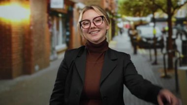 Bir kadın gülümsüyor ve şehir sokaklarında barış işareti yapıyor, gözlük ve koyu renk bir ceket giyiyor, gündüzleri güven ve mutluluk saçıyor..