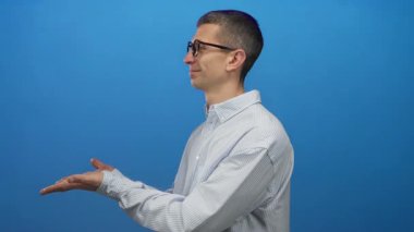 Gözlüklü bir adam mavi bir duvara gülümsüyor, çizgili bir gömlek giyiyor ve elleri açık hareket ediyor, izole bir ortamda sunum veya açıklama öneriyor.