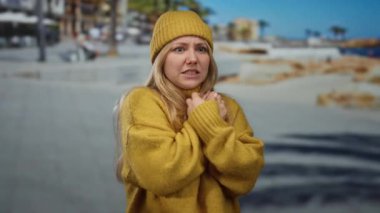 Hardal süveter ve bereli bir kadın, arka planda bulanık dalgalar ve binalarla deniz kenarında gezinirken endişeli görünüyor..