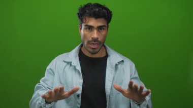 Ciddi ifadeli kot gömlek giyen İspanyol genç adam yeşil stüdyoda avuçlarını ileri doğru tutar; sakin ol..