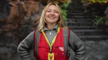 Açık havada gülümseyen, gönüllü kırmızı bir rozet takan, sarı saçlı arka planda merdivenleri çıkan, gençlik enerjisi ve toplum nişanı sergileyen bir kadın..